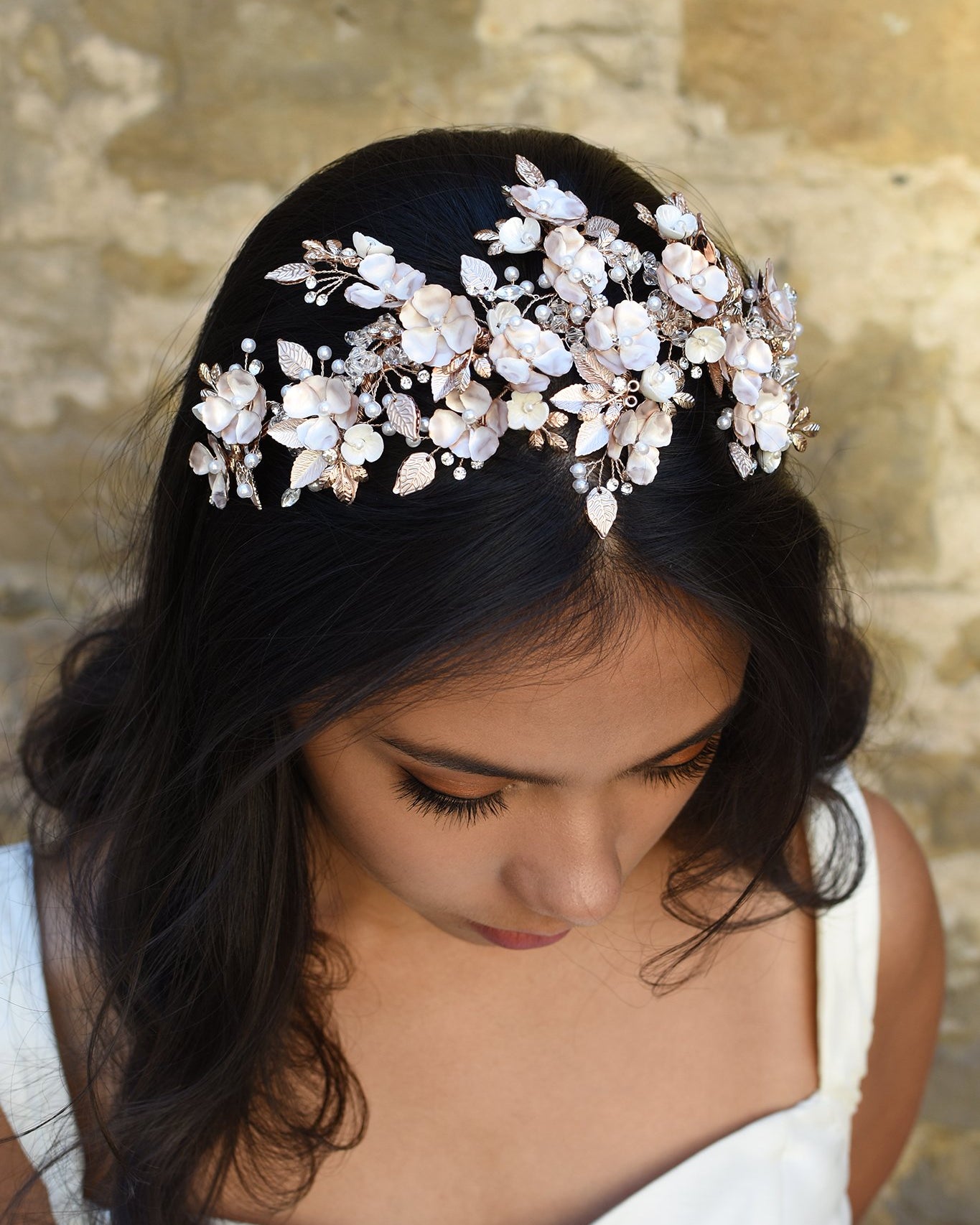 Dark Haired model wears a a pale rose gold wide headband of flowers with the background of a stone wall