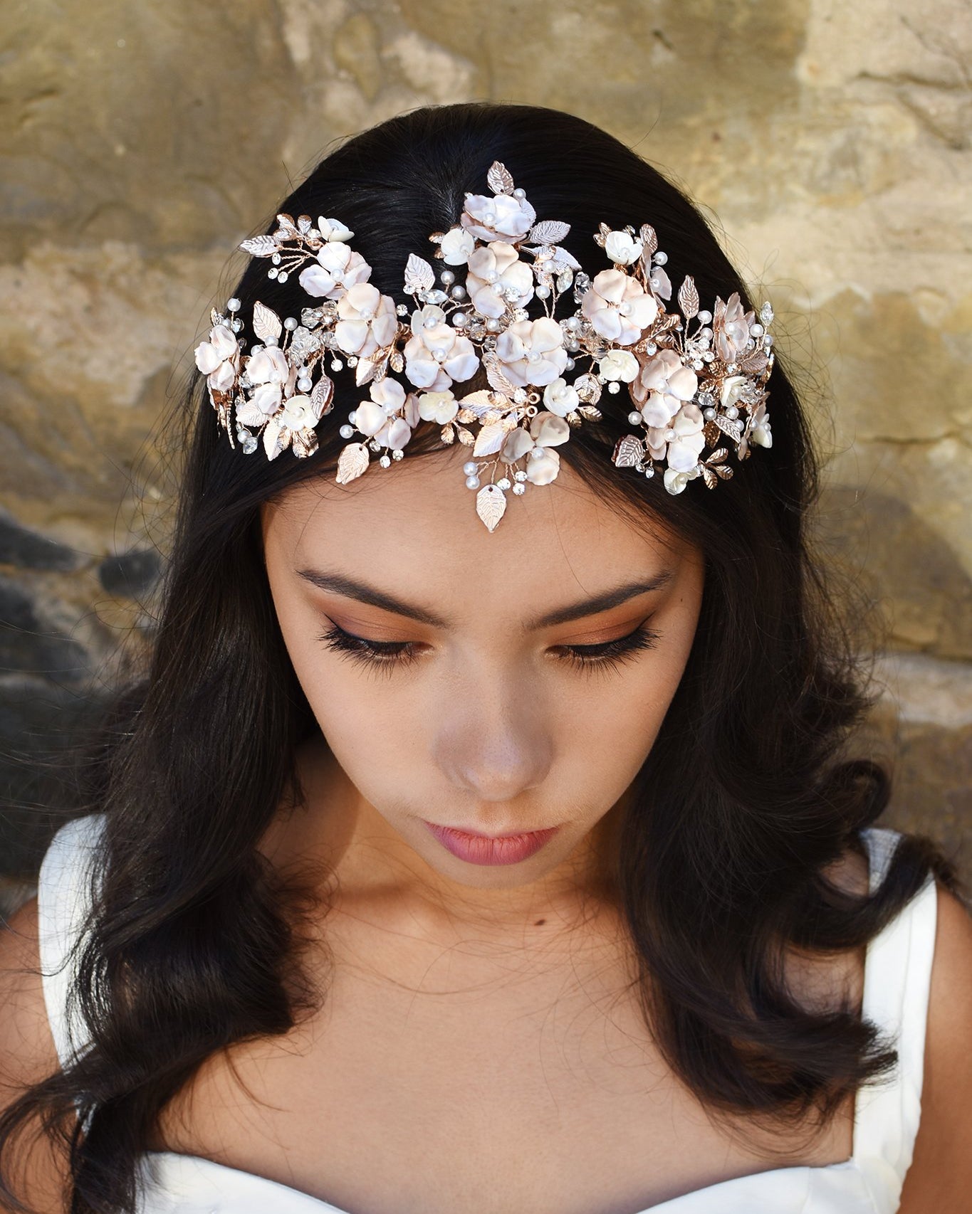Dark Haired model wears a wide rose gold headband of flowers with the background of a stone wall