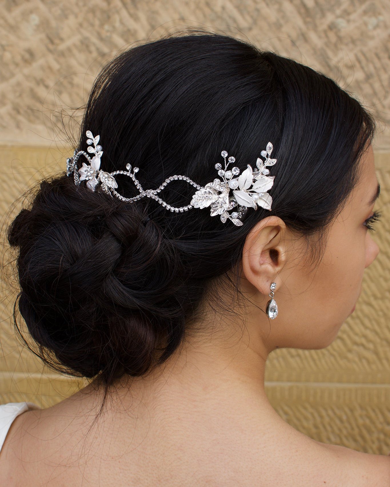 A dark haired model with curly hair wears a silver vine at the back of her head with a stone wall behind