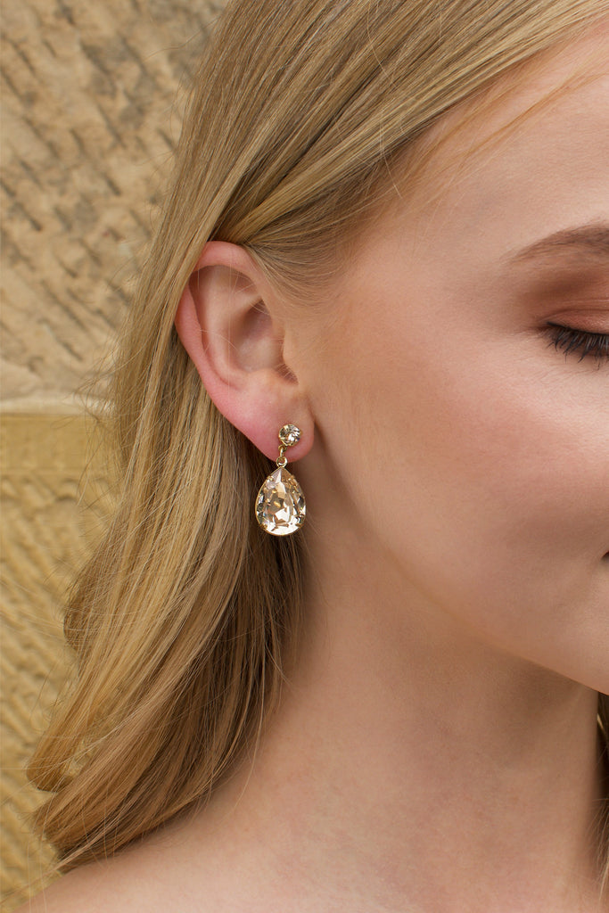 A blonde model wears a pale gold earring with a silk colour stone in her ear with a stone wall background