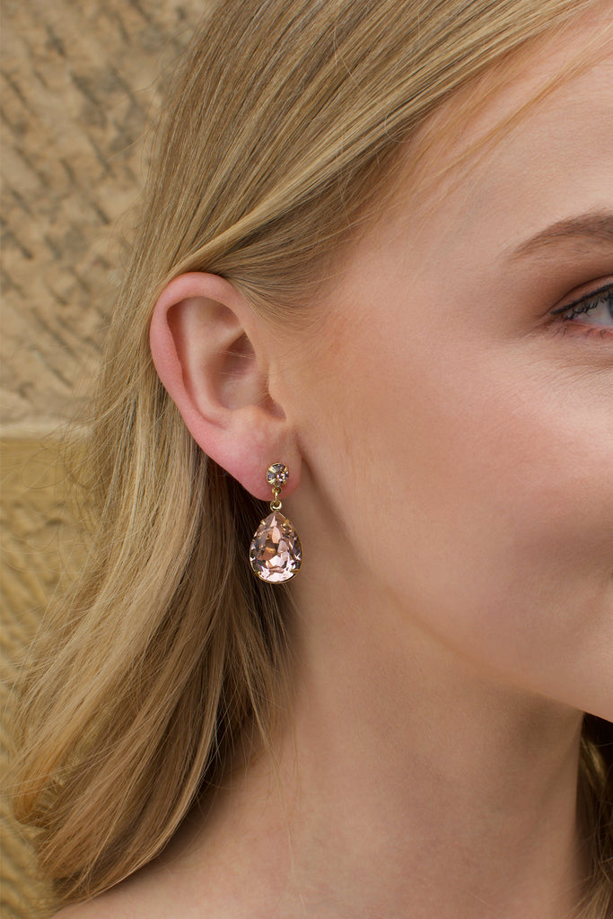 A blonde model wears a gold earring with a vintage rose colour stone with a sandstone wall background