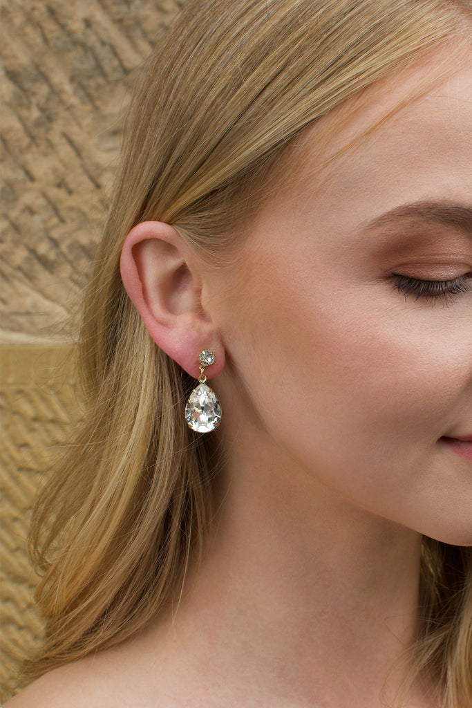 A blonde model wears a gold earring with a clear stone with a stone wall background