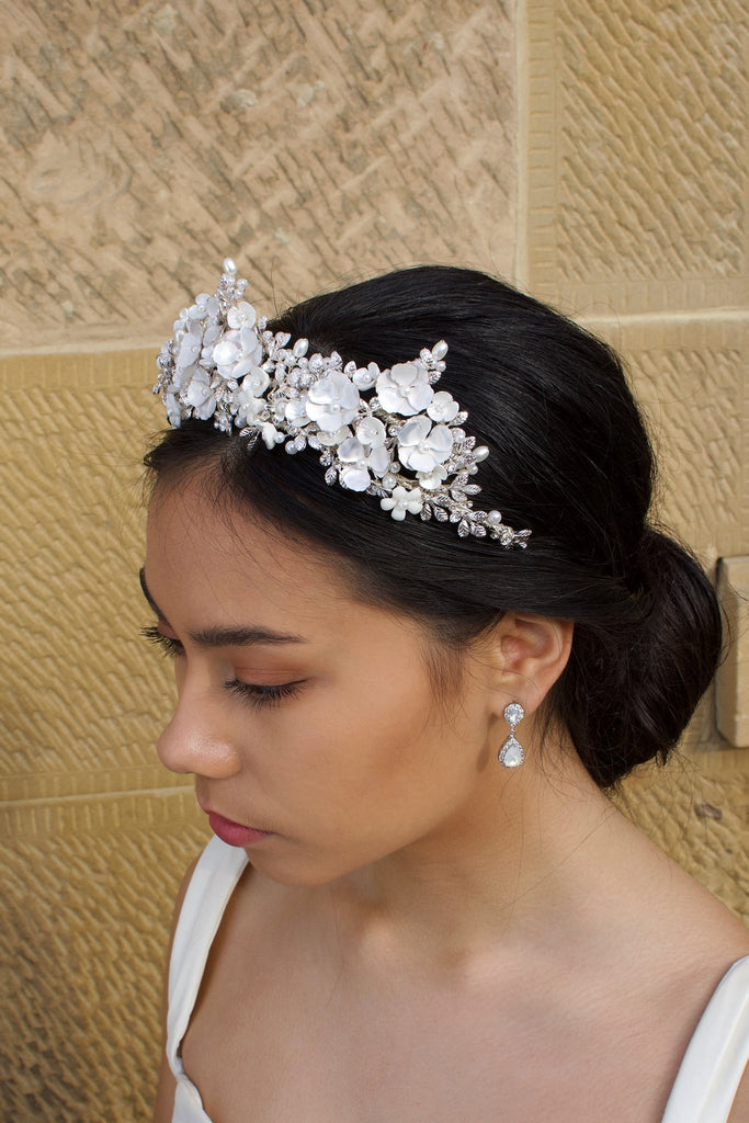 A side view of a dark haired bride wears a three pointed flower tiara at the front of her head. Behind is a sandstone wall.