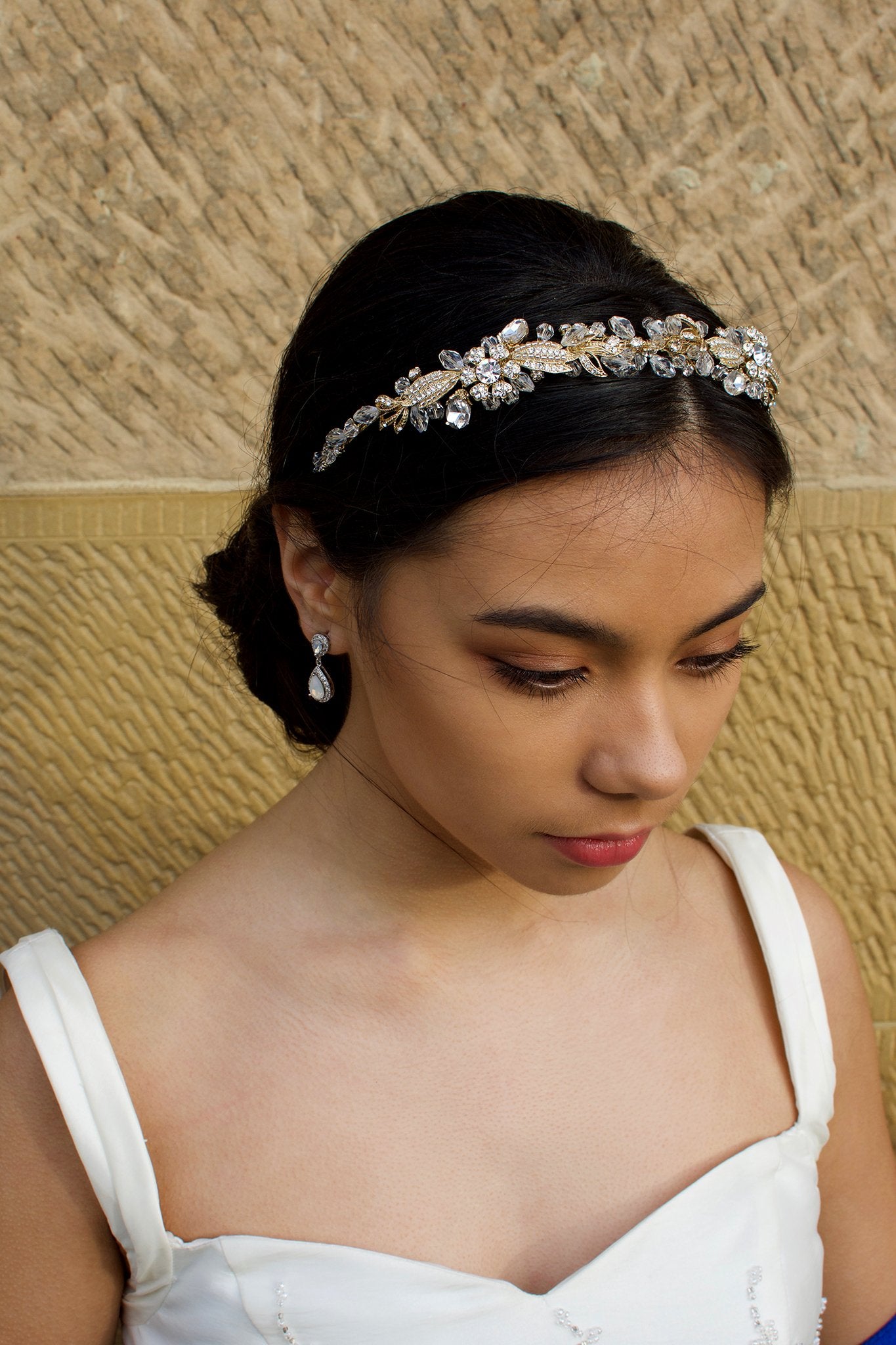 Dark haired model with long hair wears a side comb of pale gold parts at the front of her head. With a stone wall background