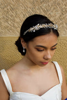 Dark haired model with long hair wears a side comb of pale gold parts at the front of her head. With a stone wall background