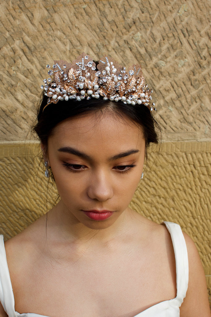 A black haired model wears a rose gold tiara with pearls in the background is a sandstone wall.