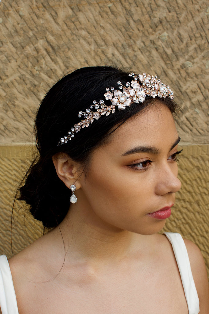 A dark haired model wears a rose gold tiara with pearl flowers against a stone wall background.