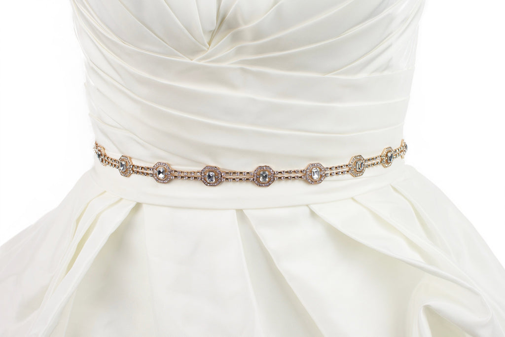 A narrow rose gold belt with crystals is fitted around the waist of an ivory bridal gown with a white background