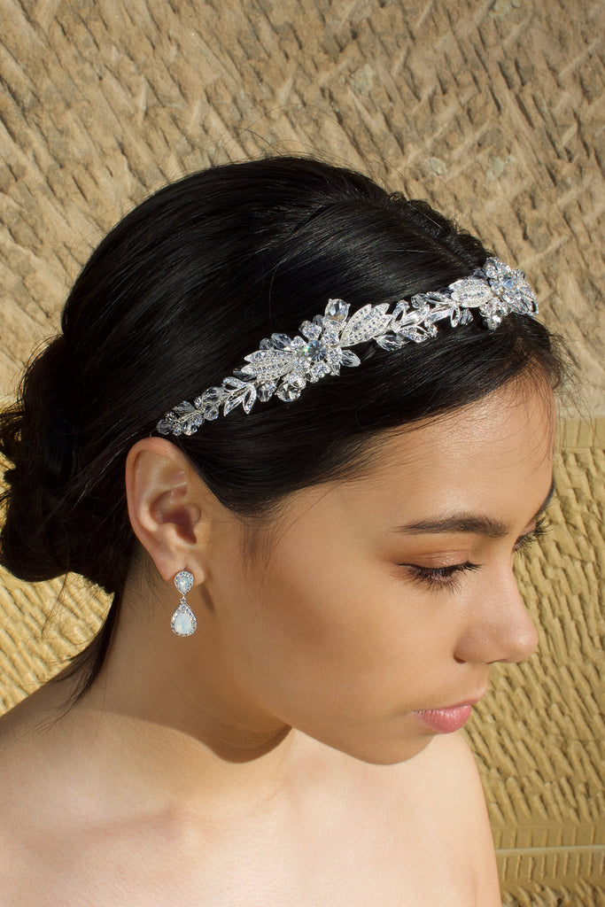 A dark haired model wears a Swarovski crystal crystal headband on the front of her head. The background is a stone wall. 