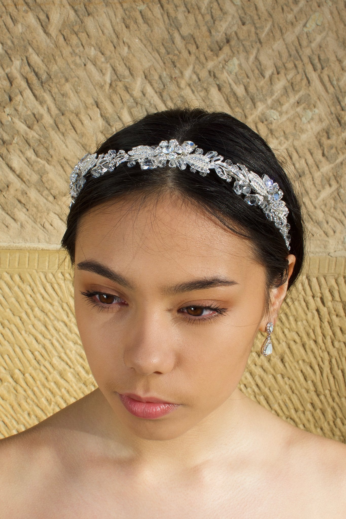 Black haired model with long hair wears a side comb of pale gold leaves at the side of her head. With a stone wall background