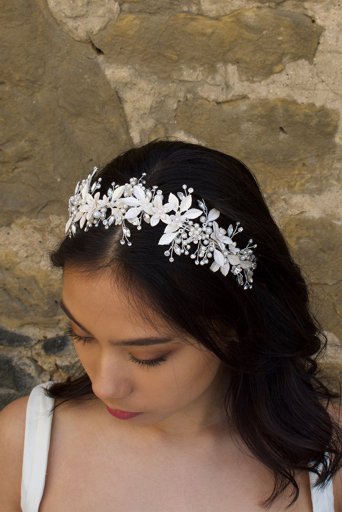 Model looks down wearing a silver leaves headband on her dark hair with an old wall behind