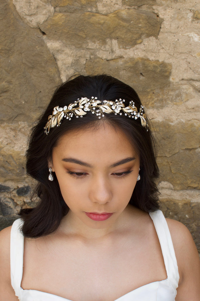 A front view of a model in front of a stone wall wearing a pale gold bridal headband on her forehead.