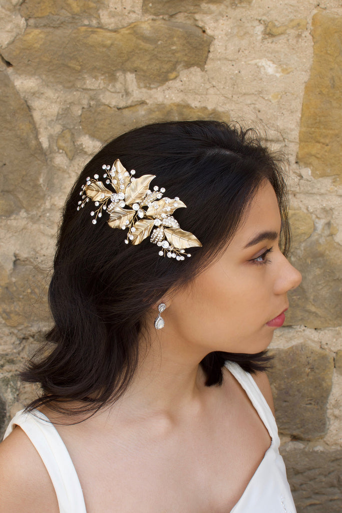 Gold leaf side comb worn by a dark hair bride standing beside a stone wall.