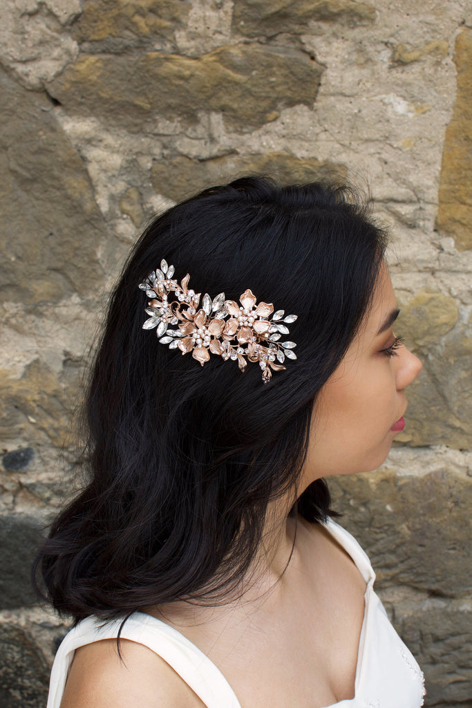 A dark haired model wears a Rose Gold flower comb on the side of her head. A stone wall is the background