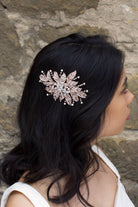 A back haired model wears a rose gold flower bridal side comb on the side of her head. A stone wall is the backdrop