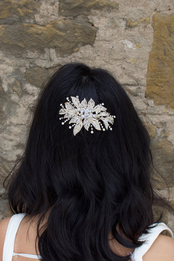 A back haired model wears a gold flower bridal side comb on the back of her head. A stone wall is the backdrop