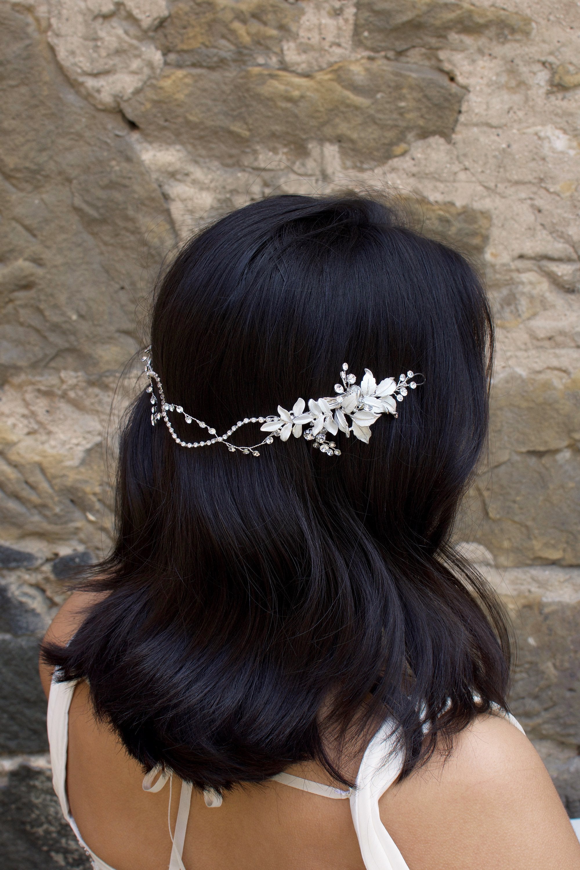 A Black haired model with her hair down wears a soft bridal vine at the back of her head. Stone wall background