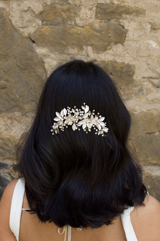 Dark haired model with long hair wears a side comb of pale gold leaves at the back of her head. With a stone wall background