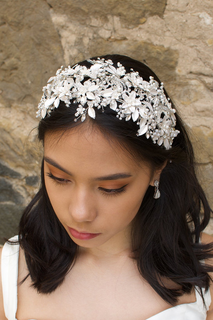 Black hair model wears a white leaves bridal headband that is wide with a stone wall behind her
