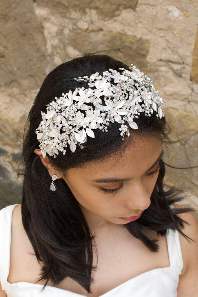 Black hair model looks down wearing a wide bridal headpiece with many leaves. Stone wall is behind