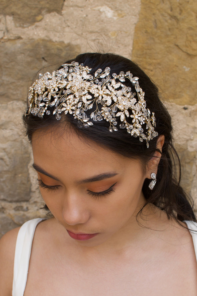 A model wears a wide golden headband across the front of her head with a stone wall background
