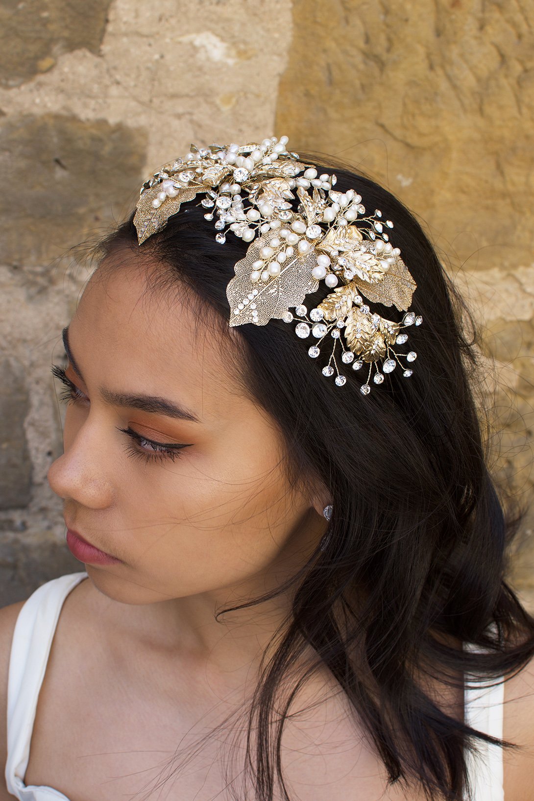 A wide gold headband worn by a dark hair bride with a stone wall background