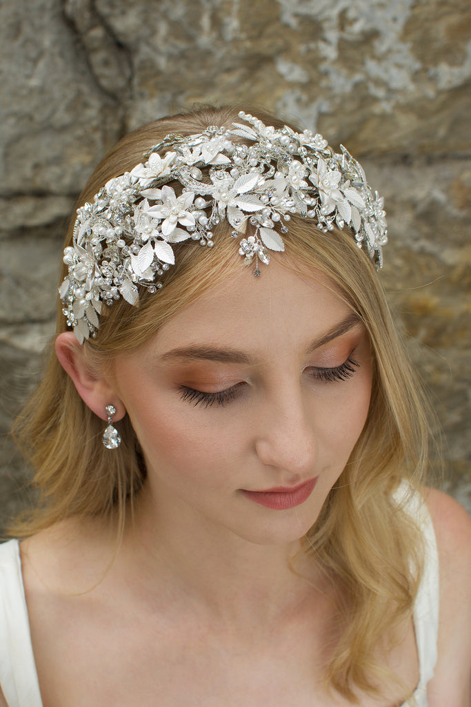 Model facing the camera wears a wide silver leaves bridal headband and the background is an old stone wall.