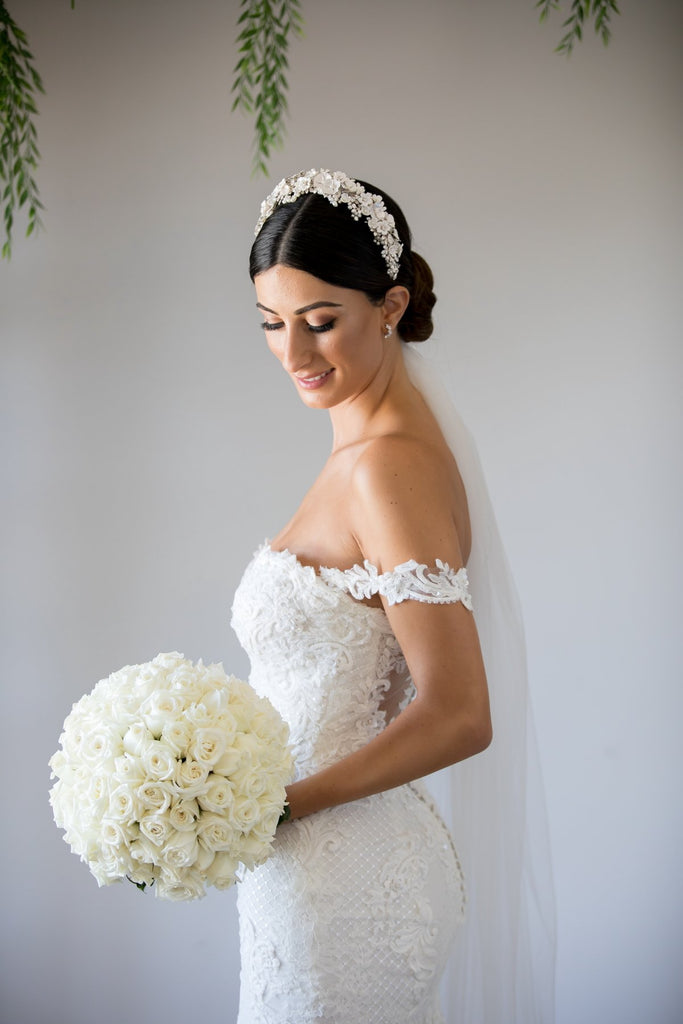 A bride wears a headband of ceramic flowers on her dark hair. There are green leaves in the background