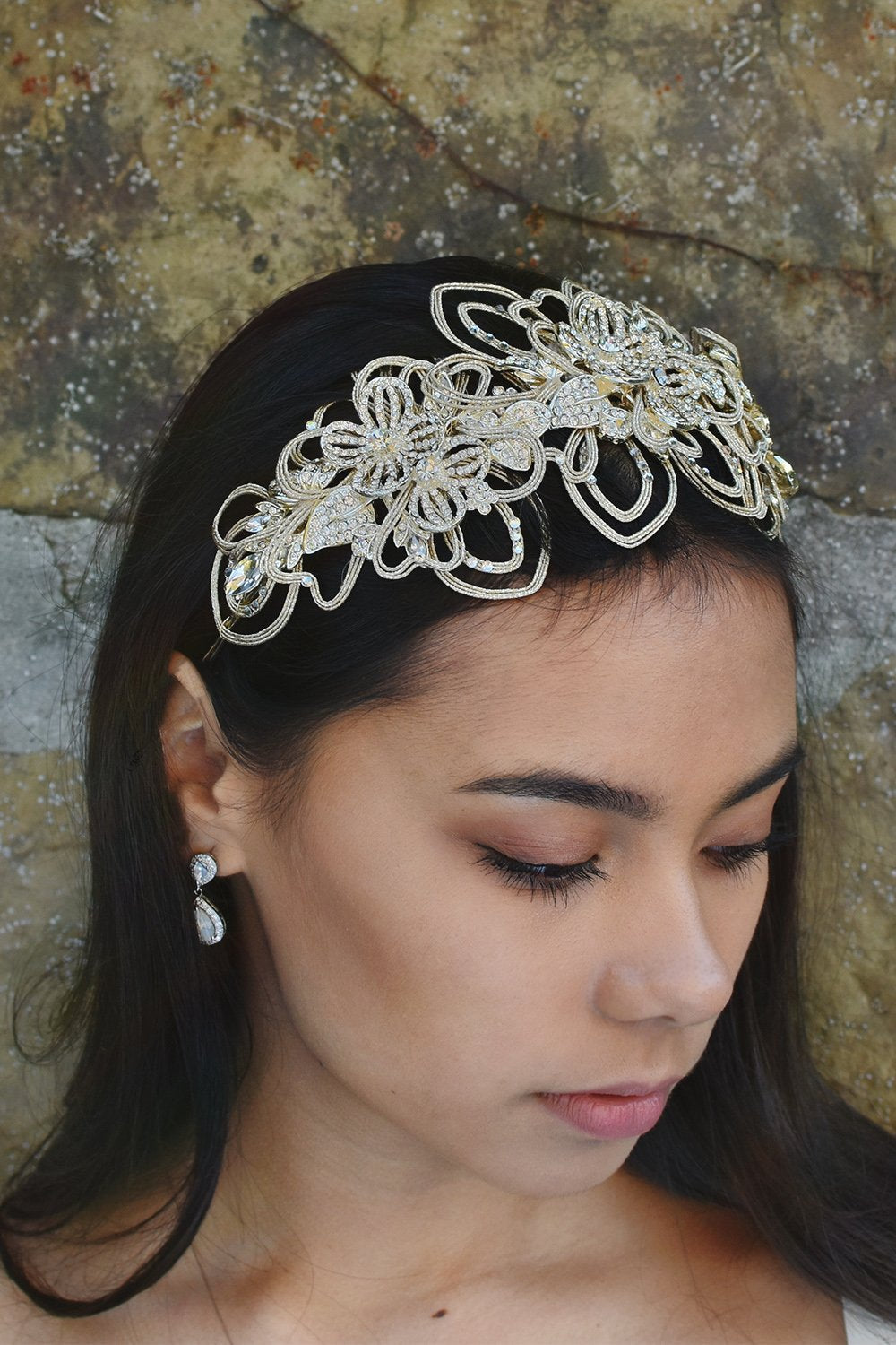 Dark haired model wearing a matt gold bridal headband at the front of her head. There is a stone wall behind her.