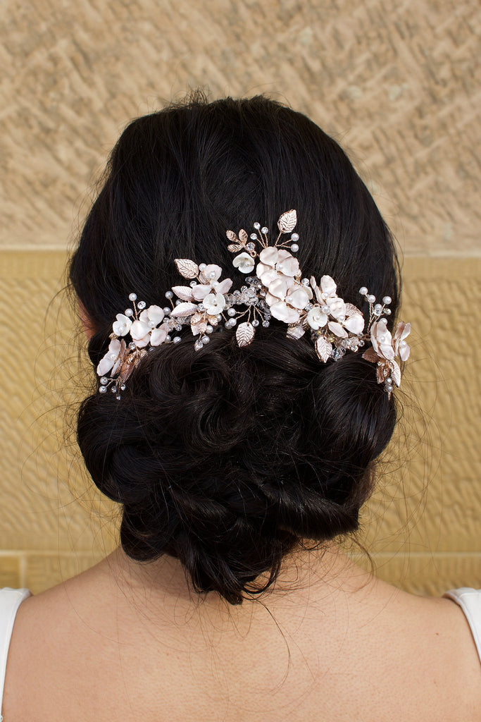 Bride with curling black hair wears a pale rose gold flowers headpiece on the back of her head. A stone wall is the background