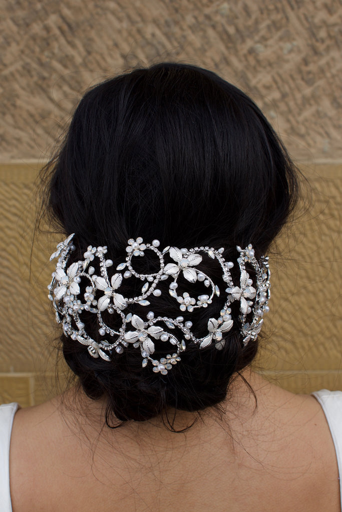 A black hair bridal model wears a wide silver hair cage on a bun of hair. There is a stone wall background