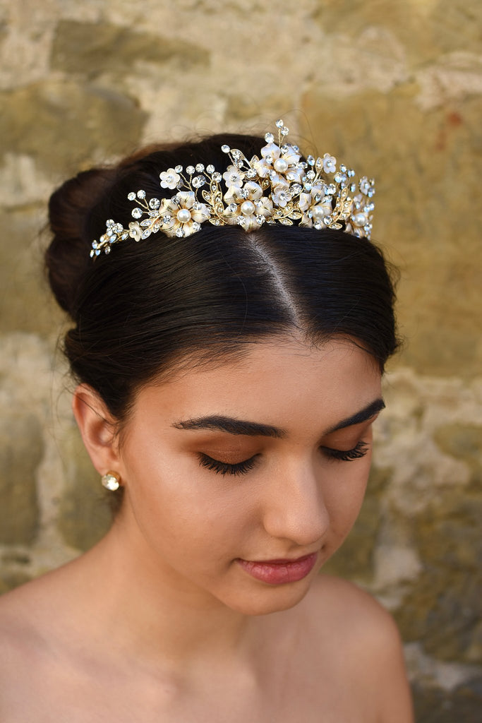 A dark  model wears a gold tiara with pearl flowers against a stone wall background.