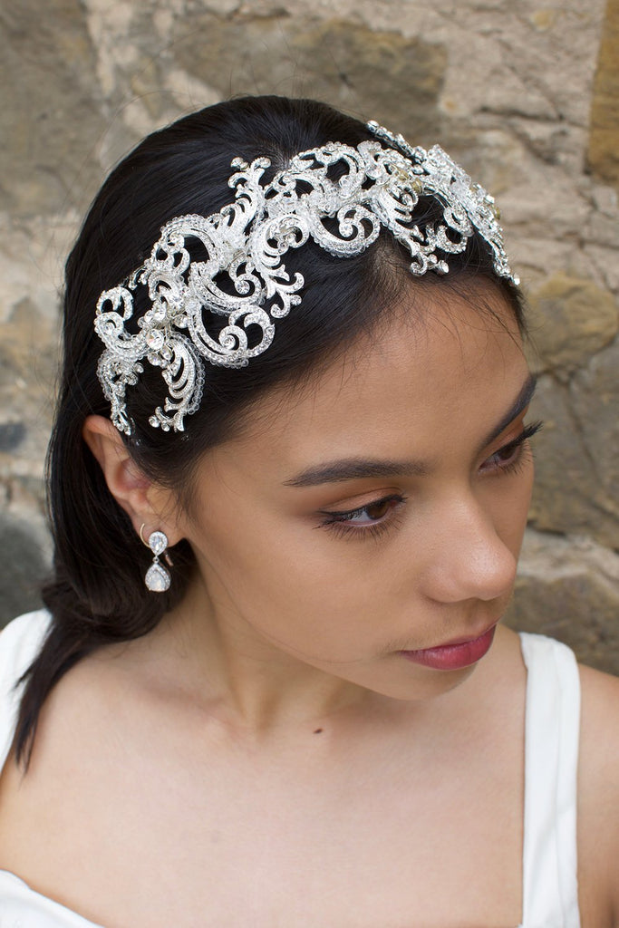 Wide swirling Bridal Headband in Silver with clear crystals worn by a dark haired model with a stone wall backdrop
