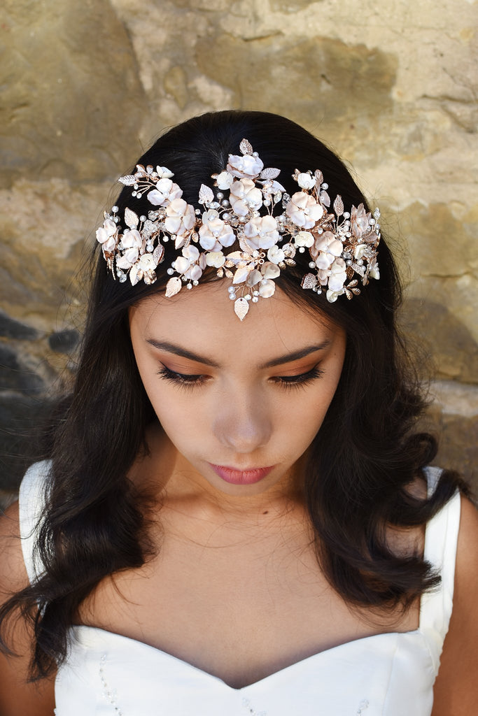Dark Haired model wears a wide rose gold headband of flowers with the background of a stone wall